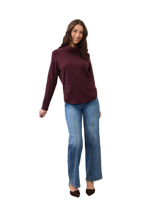 A woman modeling a dark burgundy long-sleeve turtleneck sweater with front patch pockets, paired with blue jeans and brown pointed-toe flats, standing against a white studio wall.