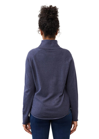 A woman viewed from the back wearing a dark slate blue mock neck long-sleeve top and dark blue jeans, set against a plain light gray studio background.