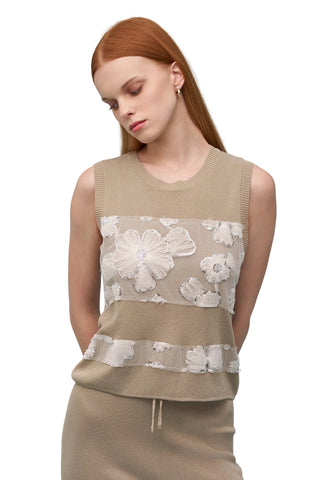 Woman wearing a beige sleeveless top with floral lace overlay against a light blue background