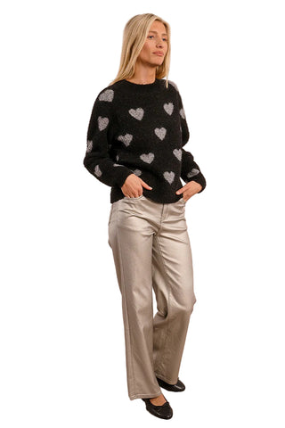 Woman wearing a black sweater with heart patterns and silver pants against a beige background