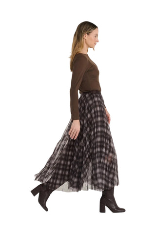 A woman in a brown long-sleeve top and a black and brown plaid pleated midi skirt walks in profile against a light gray studio background.