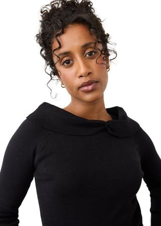 A woman with curly dark hair wearing a black long-sleeve top with a wide collar, photographed against a plain light background.