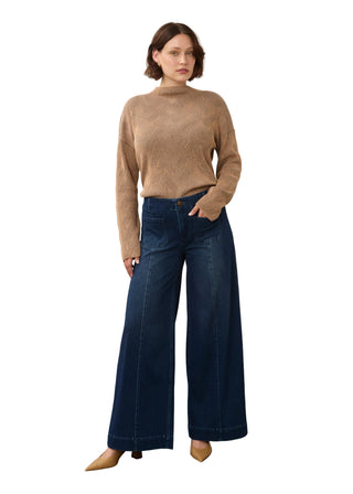 A woman modeling a beige ribbed mock-neck sweater and dark blue wide-leg jeans against a neutral studio backdrop.