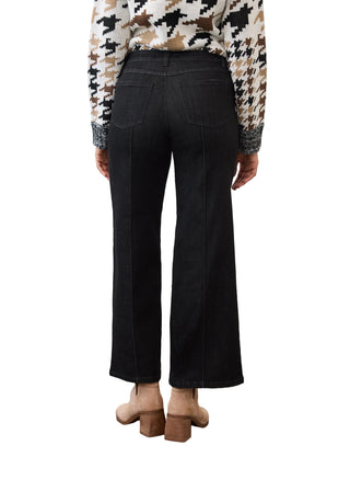 A person shown from the back wearing black wide-leg cropped jeans, a beige and black houndstooth patterned long-sleeve top, and tan block-heel mules, standing against a white wall and concrete floor.