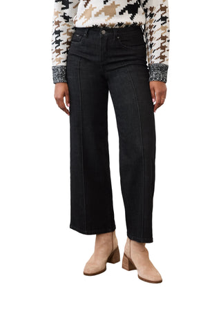 A model is wearing a pair of black wide-leg cropped jeans, paired with a patterned sweater and beige ankle boots, standing against a plain white wall and concrete floor.