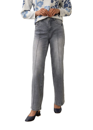 A model wearing light gray washed denim jeans with front seam detailing, paired with a blue and white patterned sweater and navy blue heels, standing against a neutral studio backdrop.