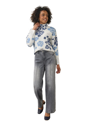 A woman modeling a white mock-neck knit sweater with a blue floral pattern, paired with wide-leg gray jeans and navy shoes, standing against a plain off-white wall.