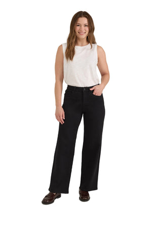 A woman modeling a pair of black wide-leg trousers with a white sleeveless top and dark brown loafers, standing against a light gray studio background.