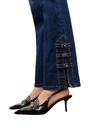 Close-up of a person wearing dark blue wide-leg jeans with decorative button details on the lower hems and black pointed slingback heels with gold hardware, shown against a light gray wall and concrete floor.