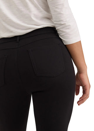 Close-up back view of a person wearing black high-waisted leggings with a back pocket, paired with a white long-sleeve top, against a light gray studio background.