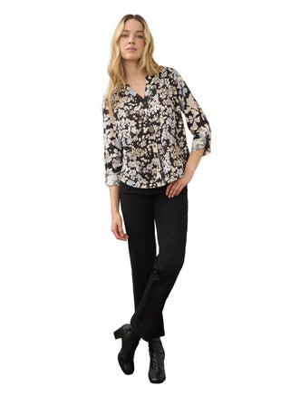 A woman modeling a black, white, and beige floral print blouse with three-quarter sleeves, paired with black trousers and black ankle boots, standing against a neutral studio backdrop.