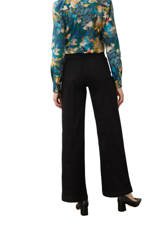 A model shown from the back wearing black wide-leg trousers and a blue, yellow, and green floral long-sleeve blouse, standing against a plain off-white wall.