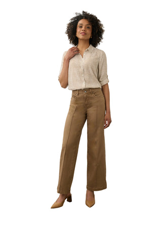 A model stands in a studio wearing a light beige textured button-up shirt, brown wide-leg cropped pants, and tan pointed-toe heels against a plain off-white wall and concrete floor.