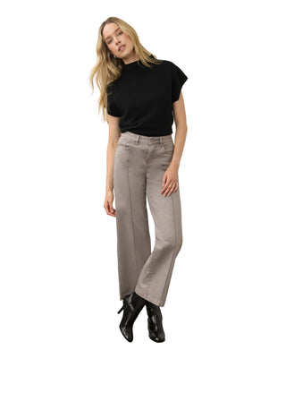 A woman modeling a black short-sleeve mock neck top and light gray wide-leg jeans with black ankle boots, standing against a white wall and concrete floor.