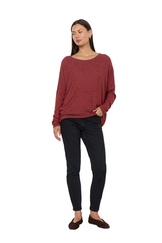 A woman modeling a loose-fitting, long-sleeve boat-neck knit sweater in a heathered burgundy red, paired with black skinny pants and dark brown slip-on shoes, standing against a plain white wall.