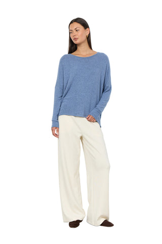 A woman modeling a heathered blue long-sleeve knit top and cream wide-leg trousers against a plain white wall.