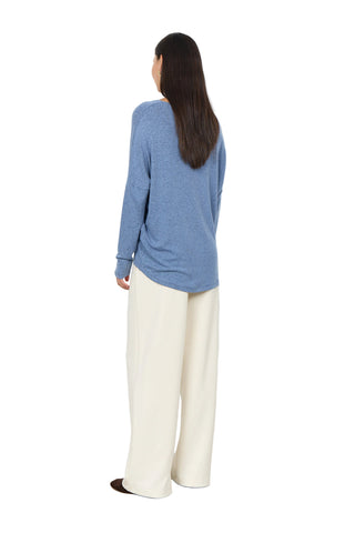 A woman is shown from the back wearing a heathered blue long-sleeve top and cream wide-leg trousers, standing against a plain white wall.