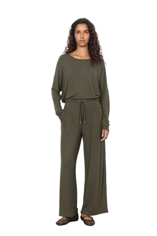 A woman modeling an olive green two-piece lounge set consisting of a long-sleeve top and matching wide-leg pants, standing against a plain white wall.