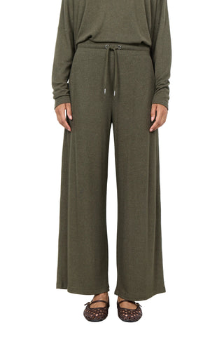 A person wearing olive green wide-leg drawstring pants with a matching long-sleeve top, paired with brown patterned flats, standing against a plain white wall.