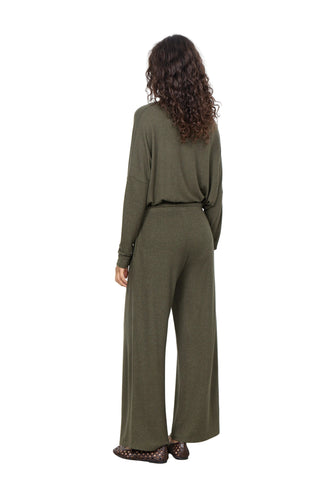 A woman with long curly brown hair is shown from behind wearing an olive green knit matching set consisting of a long-sleeve top and wide-leg trousers, paired with dark brown shoes, against a plain white wall.