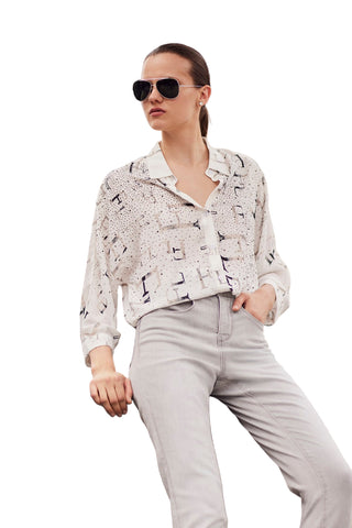 Woman wearing a patterned jacket and light pants sitting against a blurred architectural background