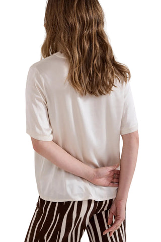 A woman viewed from behind wearing a short-sleeve cream-colored top and brown-and-white vertically striped pants, set against a plain light gray studio background.