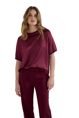 A woman modeling a matching burgundy satin short-sleeve top and trousers set against a plain light gray studio background.