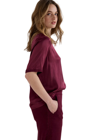 A woman wearing a matching burgundy short-sleeve top and trousers set, photographed from the side against a plain light gray studio background.
