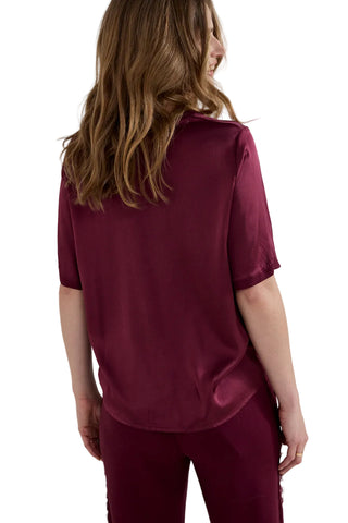A woman shown from the back wearing a short-sleeve satin-like top and matching pants in a deep burgundy or maroon color, against a plain light gray studio background.