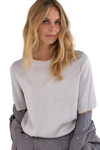 A woman with blonde hair wearing a light gray short-sleeve crewneck top, with a gray button-up shirt draped off her shoulders, against a plain light gray background.