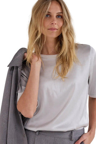 A woman with blonde hair wearing a light gray short-sleeve crewneck t-shirt and gray trousers, holding a gray button-up shirt over her shoulder against a plain light gray background.