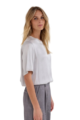 A woman with blonde hair is shown in profile wearing a white short-sleeve crewneck top and gray trousers against a plain light gray background.