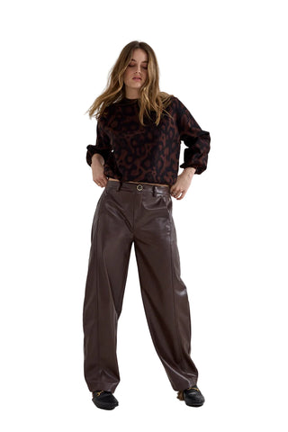 A woman modeling a dark brown leopard-print long-sleeve blouse and matching dark brown faux-leather wide-leg trousers, standing against a plain light gray studio background.