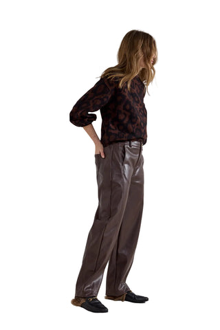 A woman stands in profile wearing a dark patterned blouse and shiny dark brown wide-leg trousers, paired with dark loafers, against a plain light gray studio background.