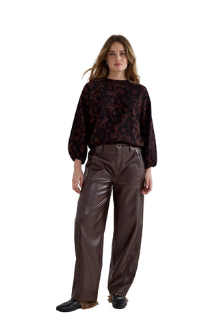 A woman stands in a studio wearing a dark brown and black patterned long-sleeve top with wide-leg brown faux-leather pants and black shoes.