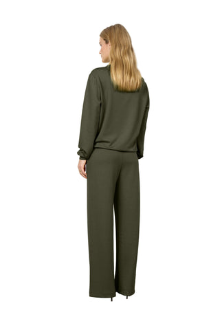 A woman with blonde hair is shown from the back wearing an olive green long-sleeve top and matching wide-leg trousers, standing against a plain white wall.