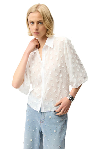 Woman wearing a white blouse with a subtle pattern and light blue pants on a plain background