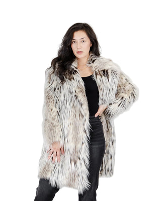 A woman modeling a long, shaggy faux-fur coat with a mottled gray, white, and black pattern, worn over a black top and black jeans against a light gray studio background.