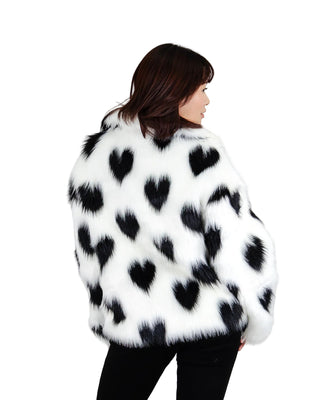 A woman shown from the back wearing a white faux fur jacket with black heart patterns, paired with black pants, against a solid light gray background.