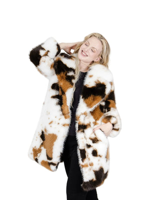 A woman modeling a long faux fur coat with a brown, black, and white cow print pattern, paired with a black top and black pants, against a solid light gray background.