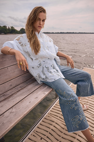 Woman sitting on a dock by a lake wearing a white embroidered top and blue jeans.
