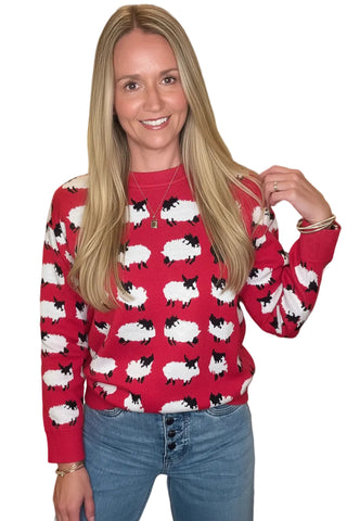A woman wearing a red crewneck sweater with an all-over black and white sheep pattern, paired with blue jeans, standing in a softly blurred indoor hallway.