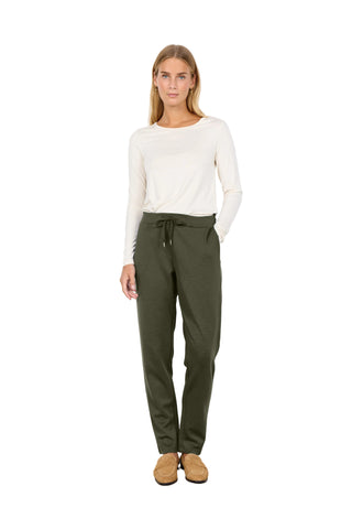 A woman stands against a plain white wall wearing olive green drawstring trousers, a cream long-sleeve top, and tan slip-on shoes.