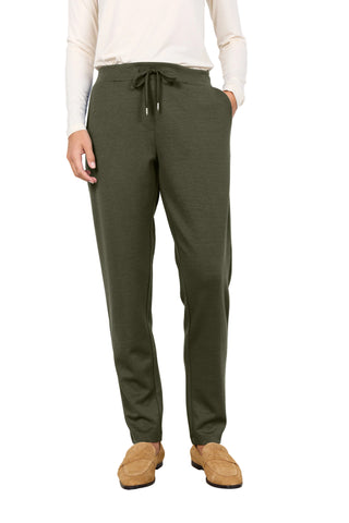 A person wearing olive green drawstring sweatpants with a cream long-sleeve top and tan suede loafers, standing against a plain light gray wall.