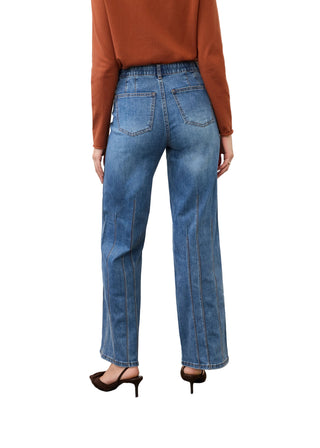 A model viewed from the back wearing high-waisted, wide-leg blue denim jeans, a rust-orange long-sleeve top, and dark brown pointed-toe heels against a white studio wall and concrete floor.