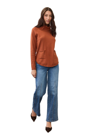 A woman modeling a rust-orange long-sleeve mock neck top with front patch pockets, paired with blue wide-leg jeans and brown pointed-toe heels, standing against a plain light gray wall.