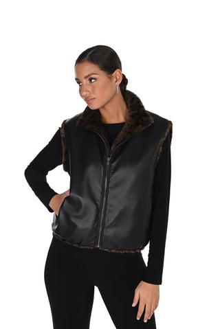 A woman modeling a black faux-leather vest with a brown faux-fur collar and hem trim, worn over a black long-sleeve top and black pants against a white studio background.