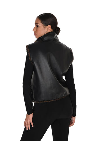 A woman shown from the back wearing a black faux-leather sleeveless vest with brown fur trim over a black long-sleeve top and black pants, against a plain white studio background.