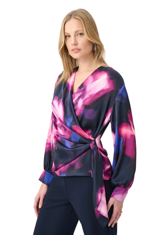 A woman modeling a long-sleeve wrap-style blouse with a dark navy base and a vibrant pink, purple, and blue abstract floral print, paired with dark pants against a light gray studio background.