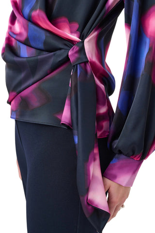 A close-up of a model wearing a long-sleeve satin blouse with a dark navy base and a vibrant magenta and blue abstract floral print, tied at the waist.